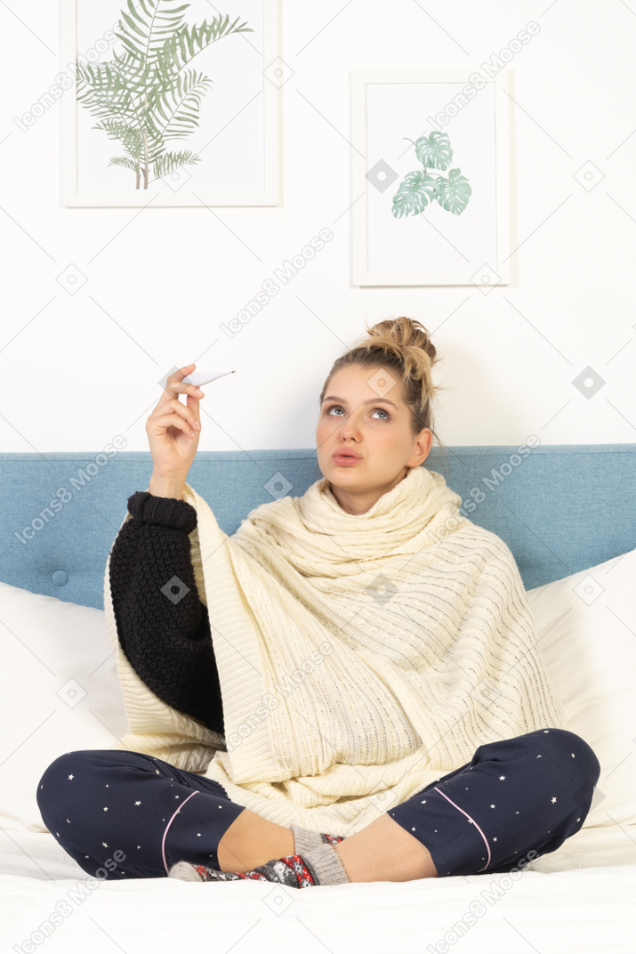 Front view of a young lady wrapped in white blanket sitting in bed with thermometer