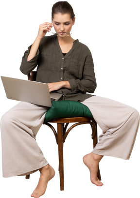 Vista frontal de una mujer joven vestida con ropa de casa sentada en una silla con una computadora portátil y poniéndose gafas