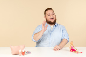 Riendo hombre grande sentado a la mesa con cosméticos y muñeca barbie