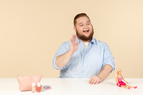 Rire grand homme assis à la table avec des produits cosmétiques et poupée barbie sur elle