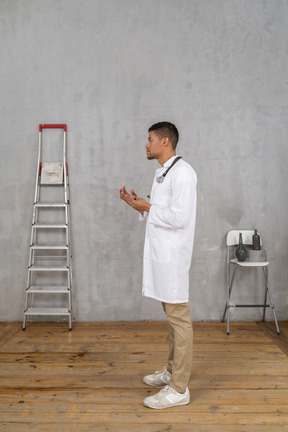 Vista lateral de un joven médico de pie en una habitación con escalera y silla explicando algo