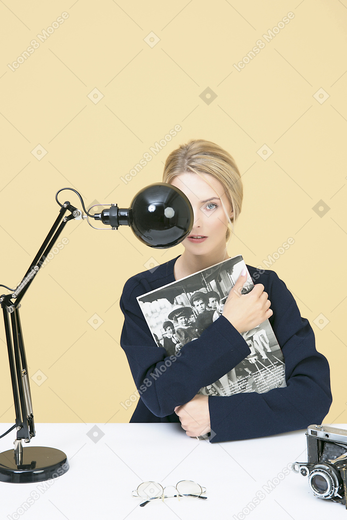 Mujer joven sosteniendo una revista y sentada en la mesa con lámpara y cámara en ella.