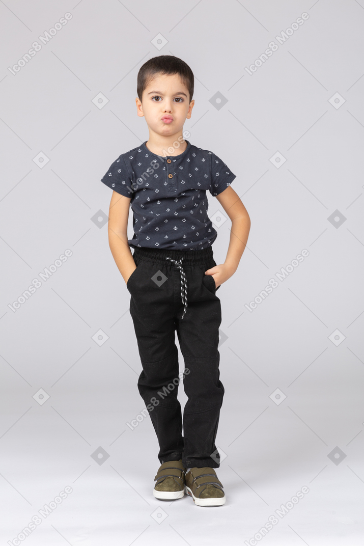 Front view of a cute boy in casual clothes posing with hands in pockets and looking at camera