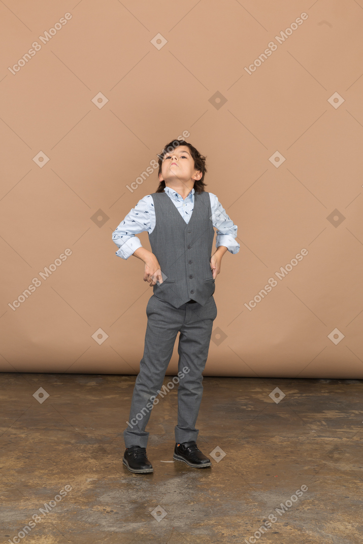 Vista frontal de um menino de terno cinza posando com a mão no quadril e olhando para cima