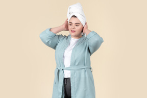 Young plus-size woman in a light blue bathrobe and with a white towel on her head, standing against a plain beige background