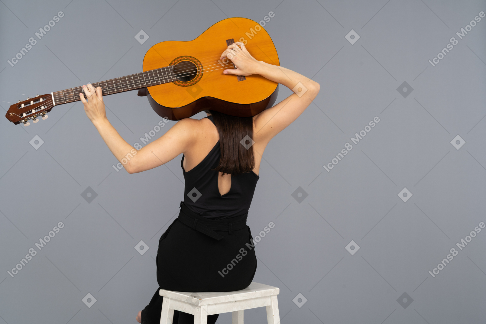 Back view of a young lady in black suit holding the guitar behind head and sitting on stool
