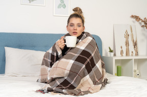 Vista frontal de una joven enferma envuelta en una manta marcada en la cama sosteniendo una taza de té