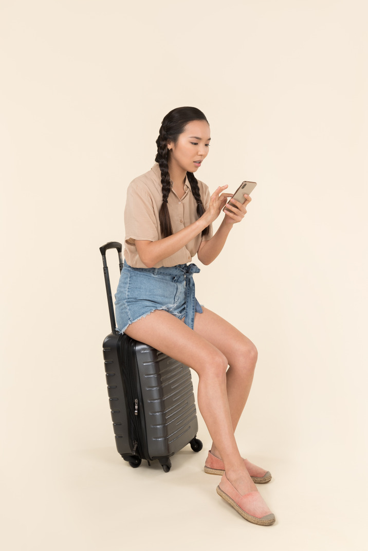 Young female traveller sitting on suitcase and using phone