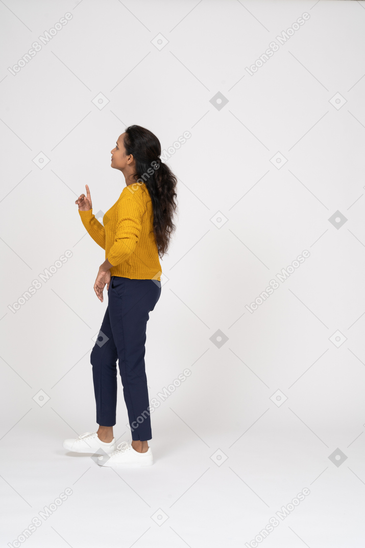 Vista lateral de una niña en ropa casual apuntando hacia arriba con el dedo