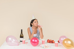 Jovem menina asiática sentado à mesa de aniversário e assobiando