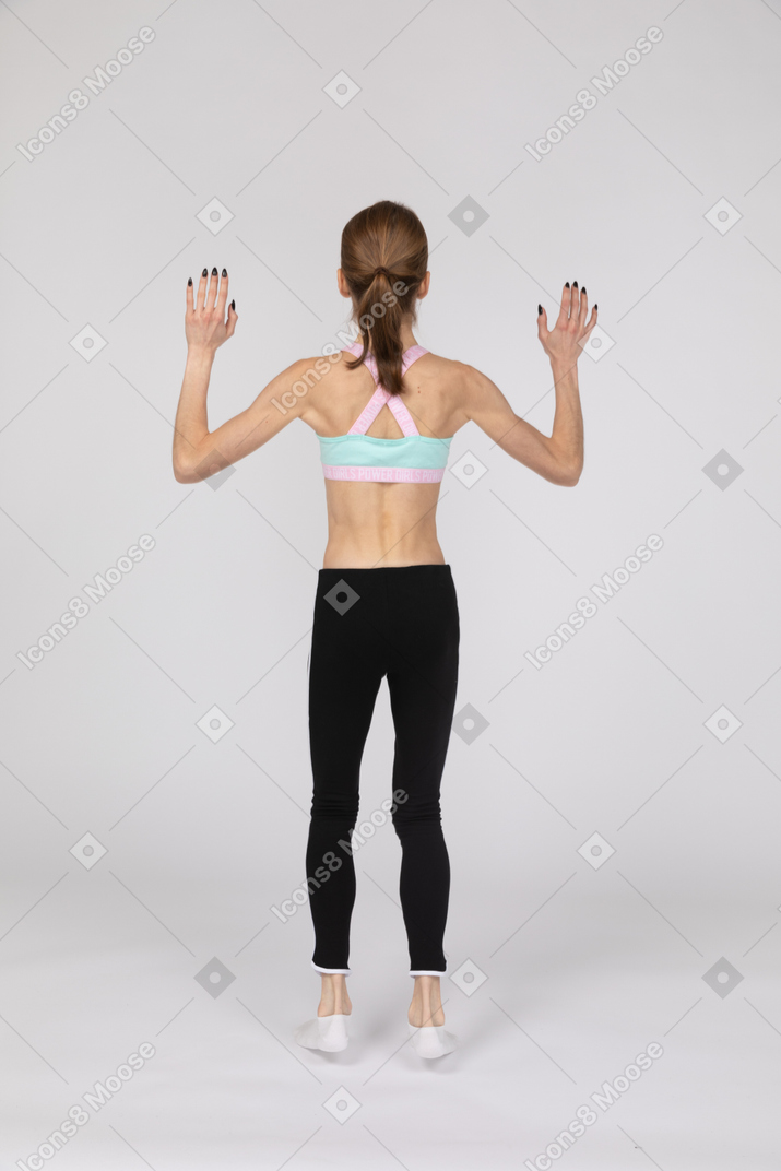 Vista traseira de uma adolescente em roupas esportivas pulando enquanto levanta as duas mãos