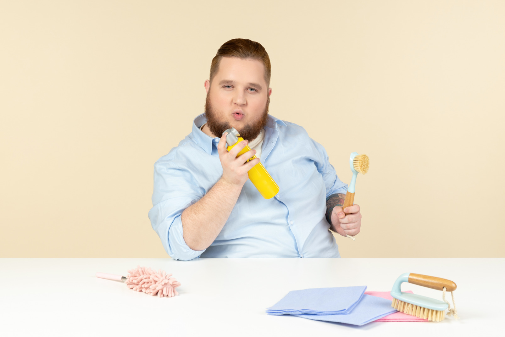 Contented young overweight man sitting at the table and holding cleaning tools