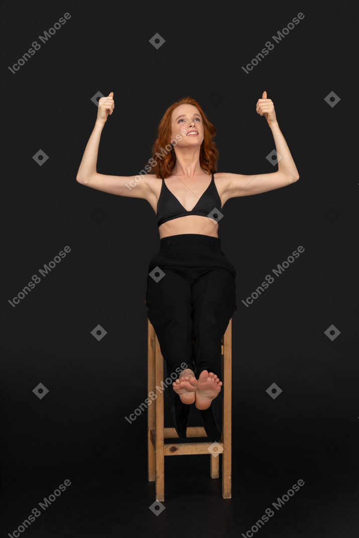 A frontal view of the beautiful woman dressed in black pants and bra, sitting on the wooden chair and holding her thumbs upher