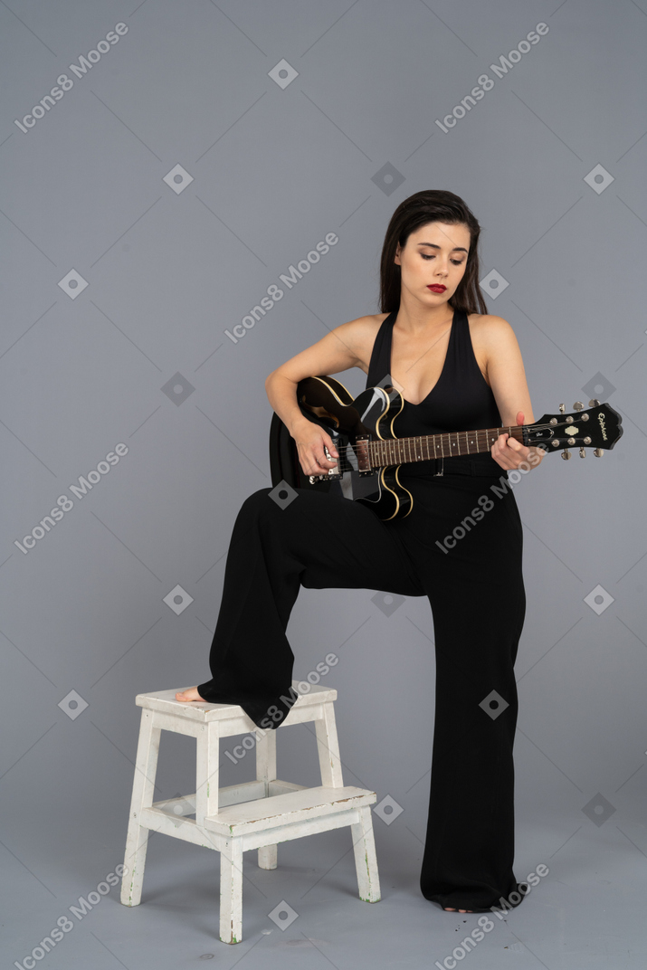 Front view of a young lady in black jumpsuit holding the guitar and putting leg on stool