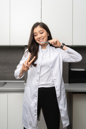 Front view of a smiling female dentist showing a peace sign and holding a toothbrush with her eyes closed