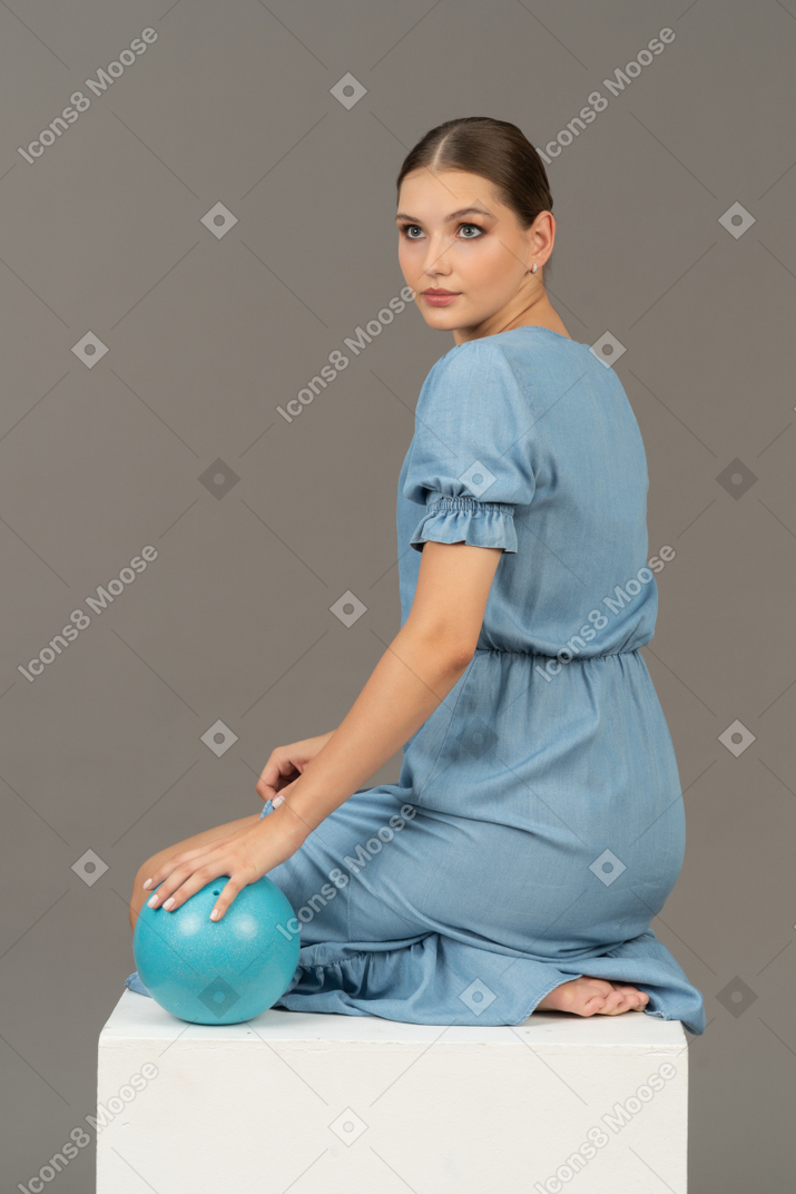 Three-quarter back view of young woman sitting on cube with blue ball