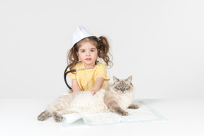 Criança menina com estetoscópio e usando chapéu médico curando um gato