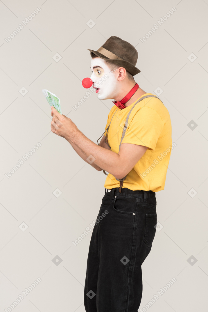 Surprised male clown standing in profile and looking at money bills he's holding
