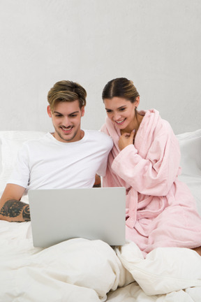 Un jeune couple heureux assis avec un ordinateur portable au lit