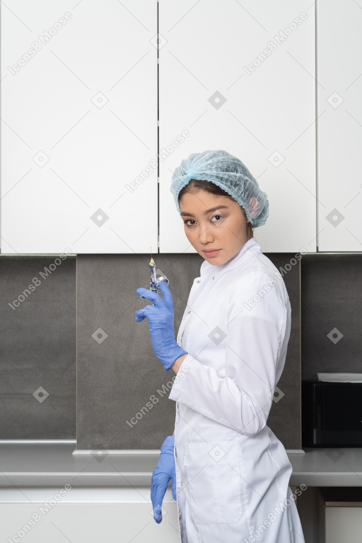 Side view of a female doctor in a medical hat holding a syringe and looking at camera