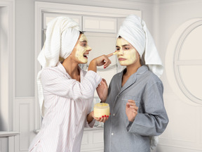 A young woman applying skin care face mask on another woman