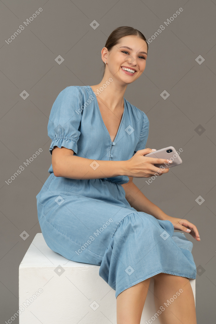 Vue de trois quarts d'une jeune femme assise sur un cube et souriant avec un smartphone à la main
