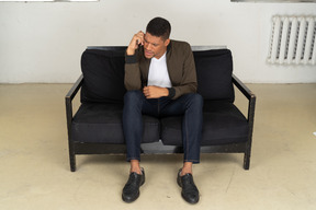 Front view of a perplexed young man sitting on a sofa and talking on his phone