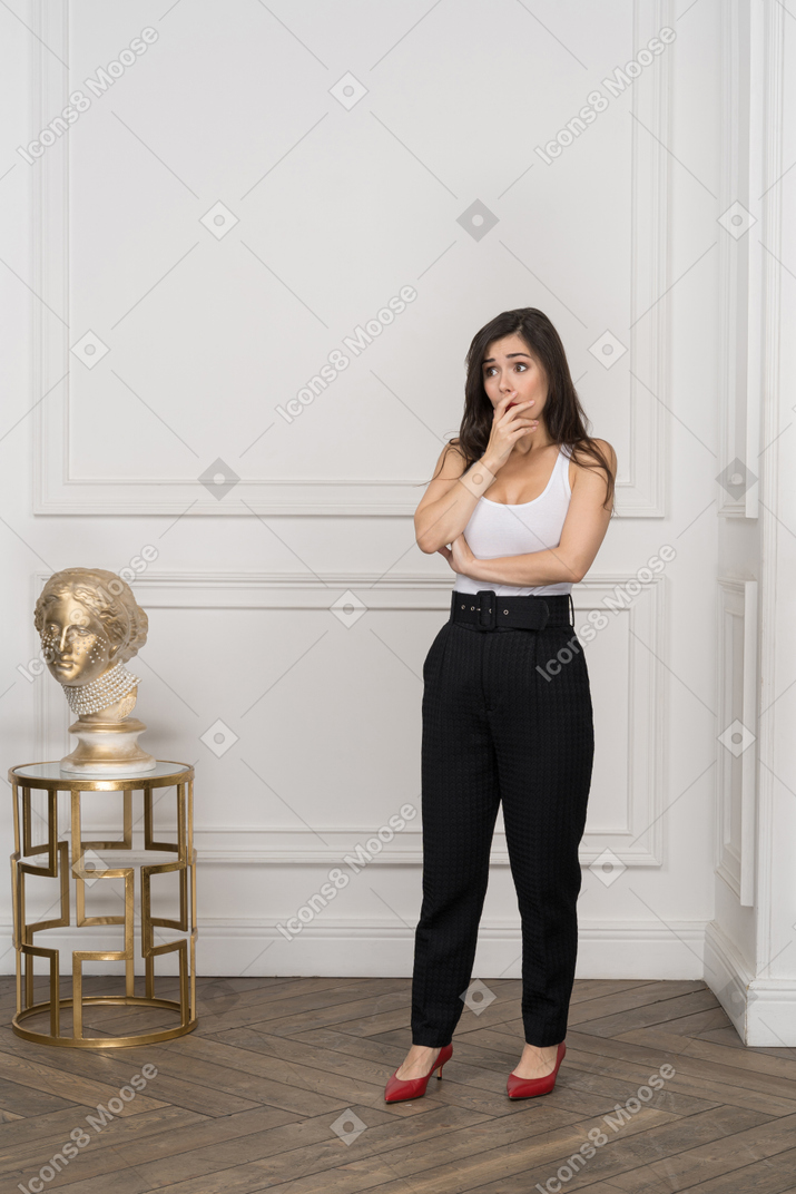 Full-length of a shocked young female touching face while standing by greek golden sculpture
