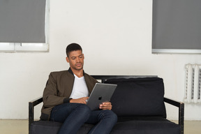 Vue de face d'un jeune homme ennuyé assis sur un canapé tout en regardant la tablette