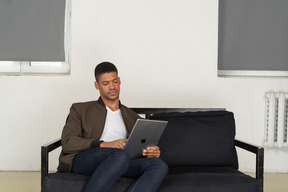 Front view of a bored young man sitting on a sofa while watching the tablet