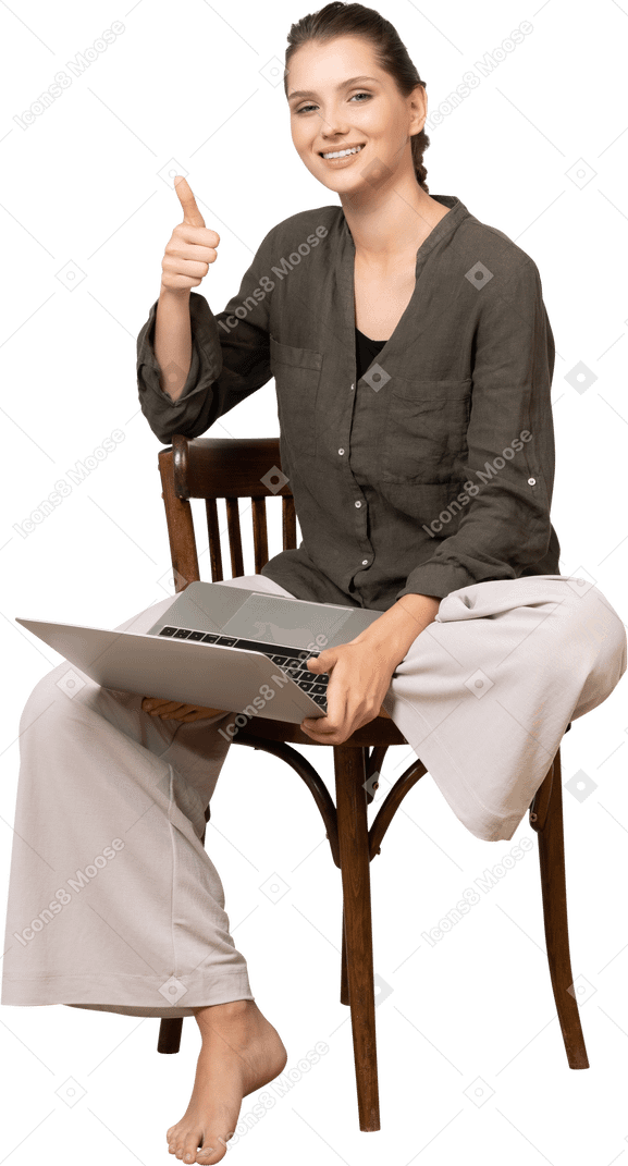 Vista frontal de una mujer joven sonriente sentada en una silla con una computadora portátil y mostrando el pulgar hacia arriba