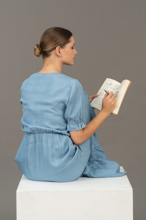 Side-back view of young woman sitting on a cube and writing in notebook
