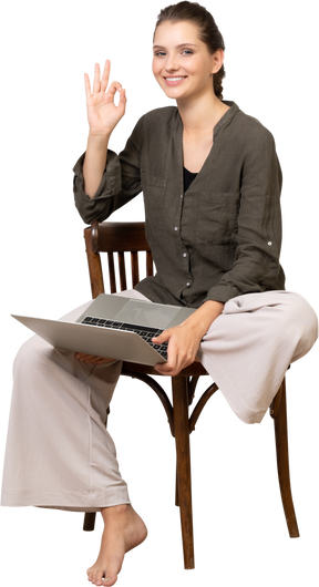 Front view of a smiling young woman sitting on a chair with a laptop & showing ok gesture