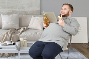 A man sitting on a chair with laptop, bottle of beer and chips in his hand