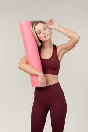Exhausted young woman in sportswear holding yoga mat