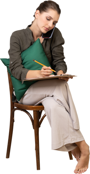 Vista frontal de una mujer joven sentada en una silla con una tableta y tener una llamada telefónica