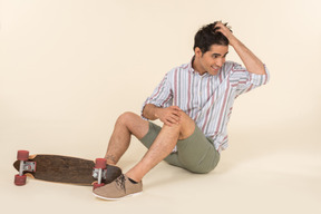 Joven caucásico ajustando el cabello y sentado cerca de skate