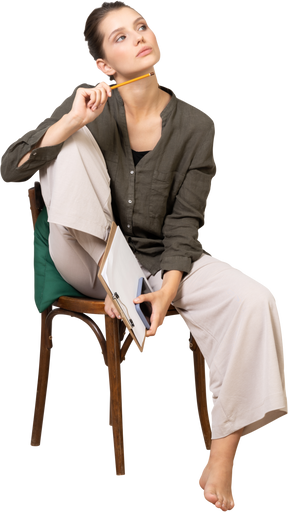 Vista frontal de uma jovem pensativa, vestindo roupas de casa, sentada em uma cadeira e fazendo anotações