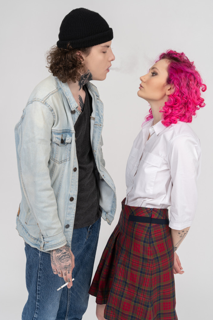 Side shot of a teen male blowing smoke to his pink-haired girlfriend