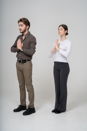 Vista de tres cuartos de una joven pareja orando en ropa de oficina tomados de la mano juntos