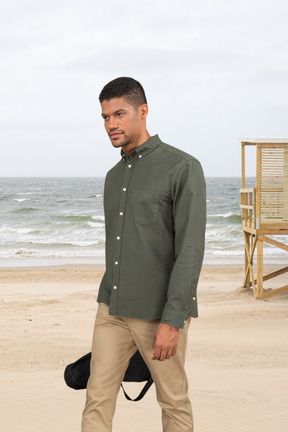 A man with a ukulele case walking on a beach in front of the ocean