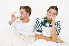 Couple sitting in bed, yawning and drinking coffee