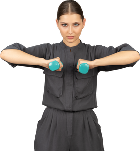Front view of young woman in a jumpsuit doing exercises with dumbbells