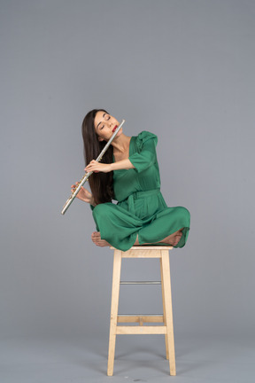 Comprimento total de uma jovem tocando clarinete sentada com as pernas cruzadas em uma cadeira de madeira