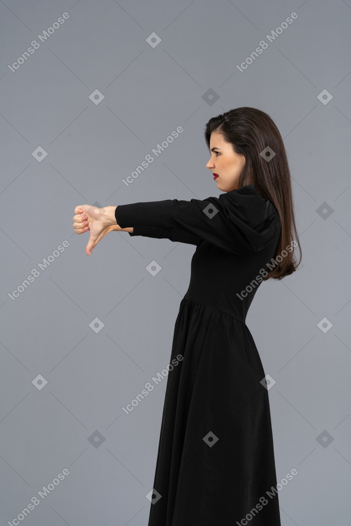 Vista laterale di una giovane donna scontenta in abito nero, mettendo i pollici verso il basso