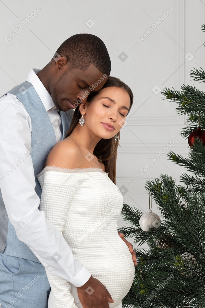 Young man and his pregnant wife standing near a christmas tree