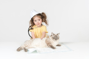 Criança menina com estetoscópio e usando chapéu médico curando um gato