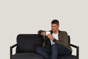 Front view of a young grimacing man sitting on a sofa with a cup of coffee