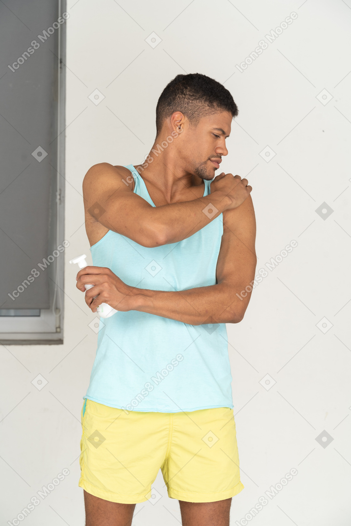 A man in a blue tank top and yellow shorts applying sunscreen