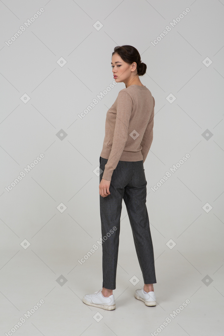 Three-quarter back view of a moody young lady standing still in pullover and pants looking aside